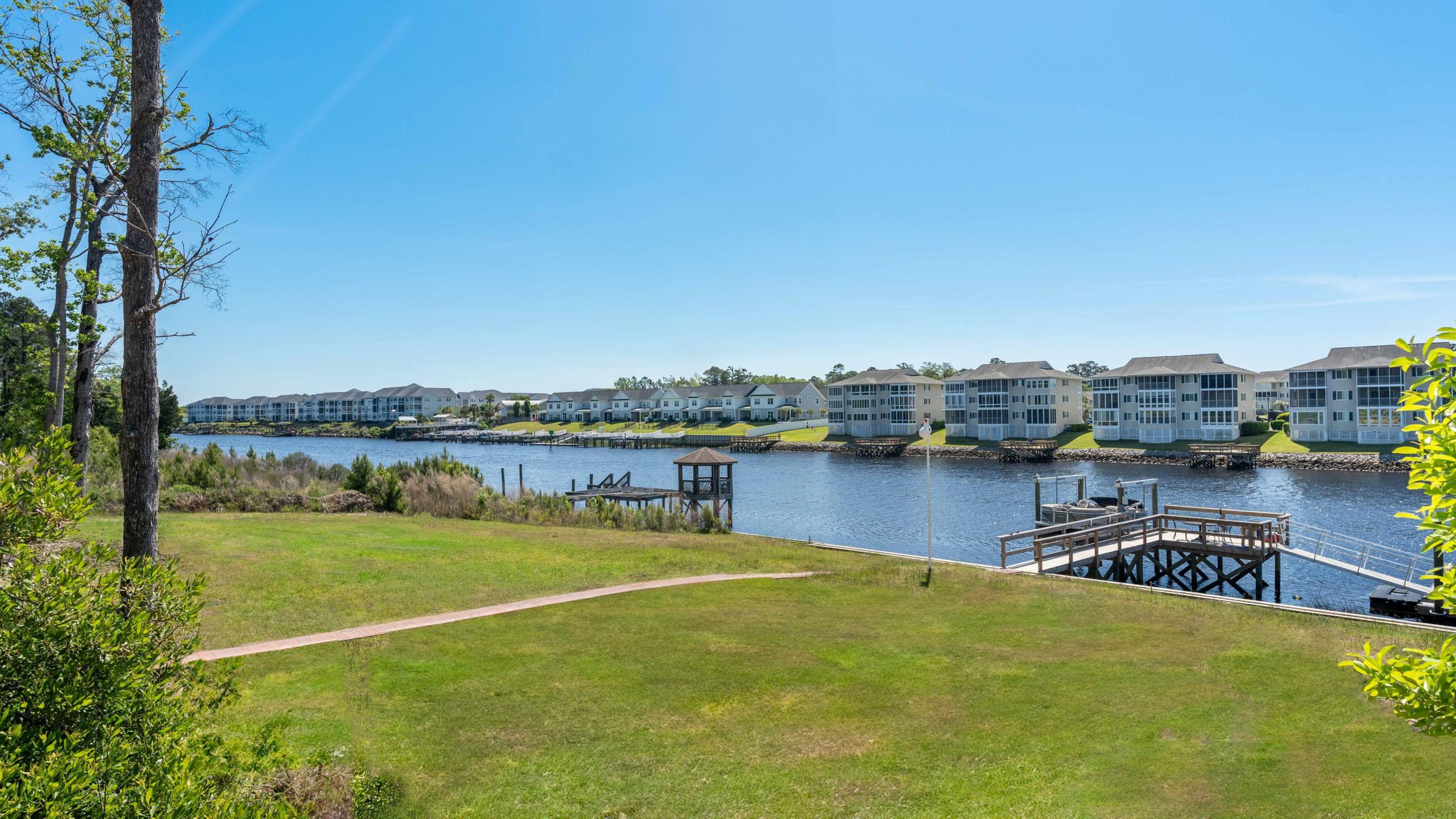 Hawthorne Waterway scene showing a grassy shoreline with a wooden pier and modern waterside homes under a bright sky.